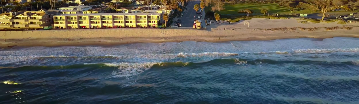 Aerial view of a Californian beach town at sunset, moving inland from the surf over palm-lined streets and homes